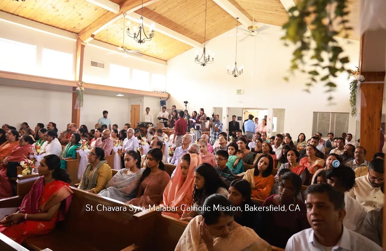 Packed congregation seated in pews, many in saris, with videographers at the rear