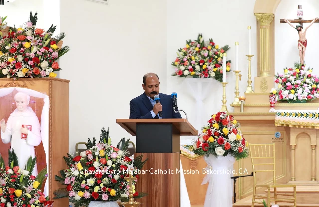 Man in a navy blazer speaking at a lectern amid large floral arrangements in the sanctuary