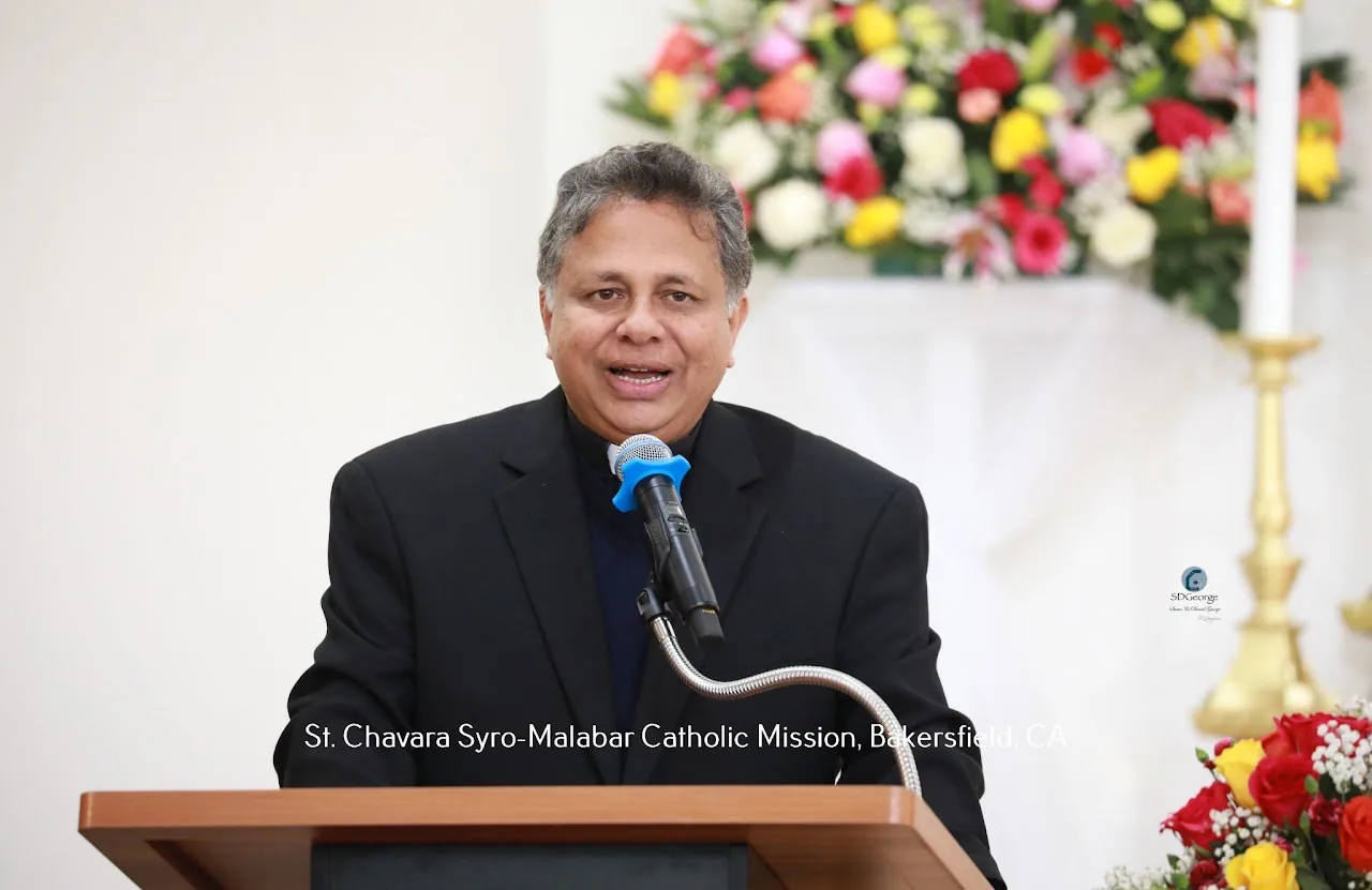 Priest in black clerical suit speaking at a wooden lectern beside flowers and a gold candlestick