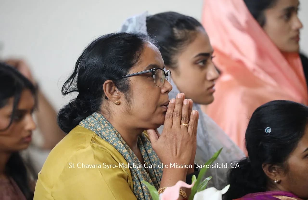 Woman in yellow kurta with hands joined in prayer during Mass, worshippers blurred behind