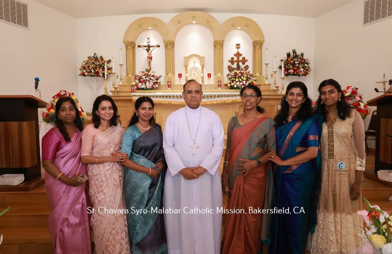 Priest in white cassock posing with six women in saris in front of the flower-decorated altar