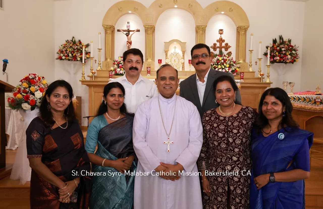 Priest in white cassock posing with six parishioners in front of the decorated altar