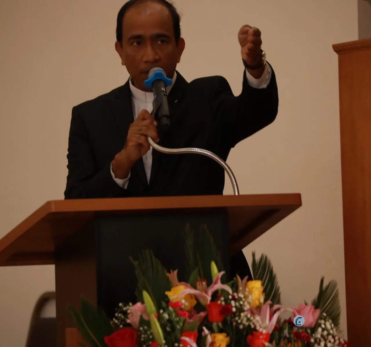 Priest speaking into a microphone at a lectern with fist raised, flowers in the foreground