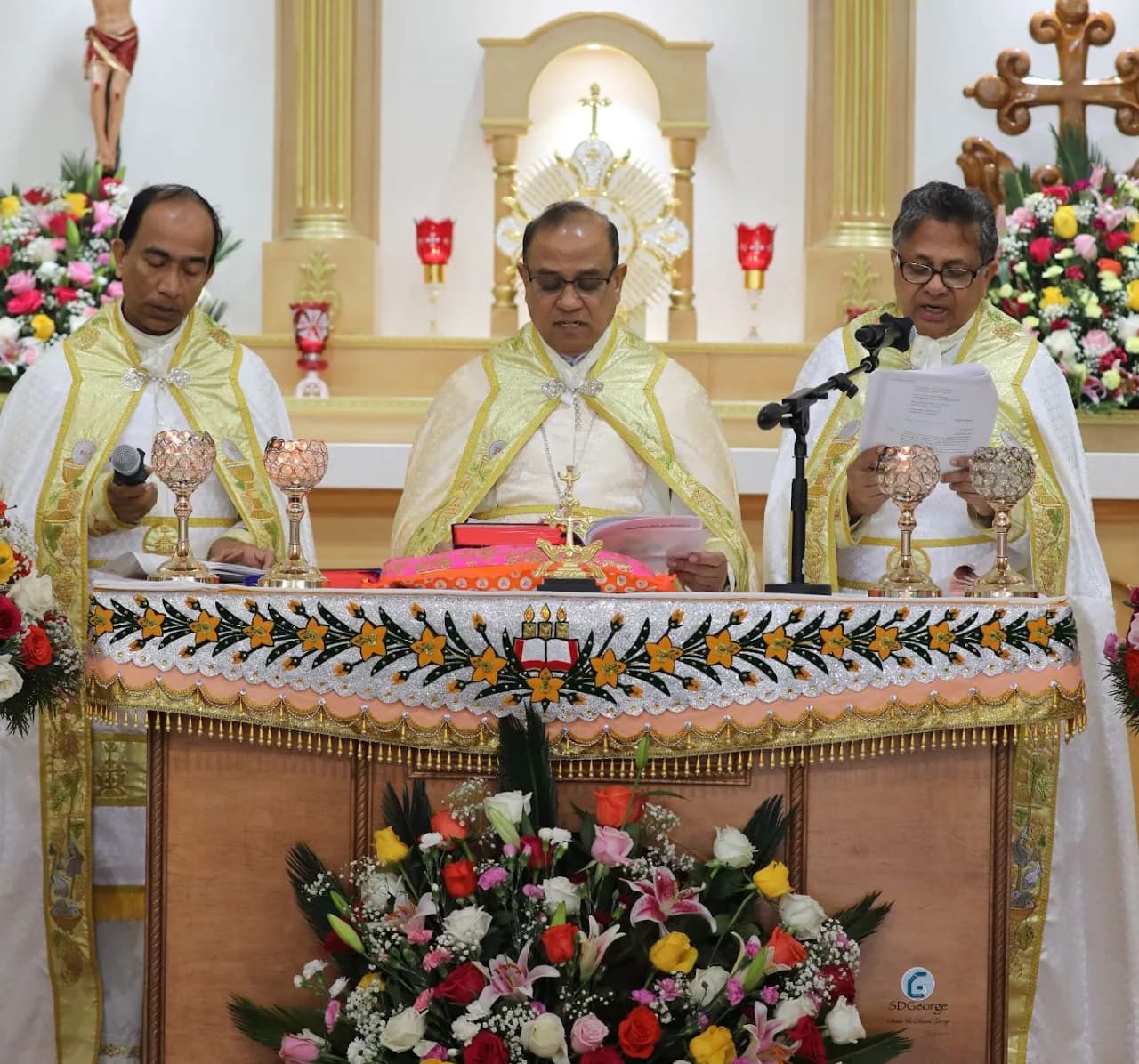 Three vested clergy at a decorated sanctuary table, one reading a document into a microphone