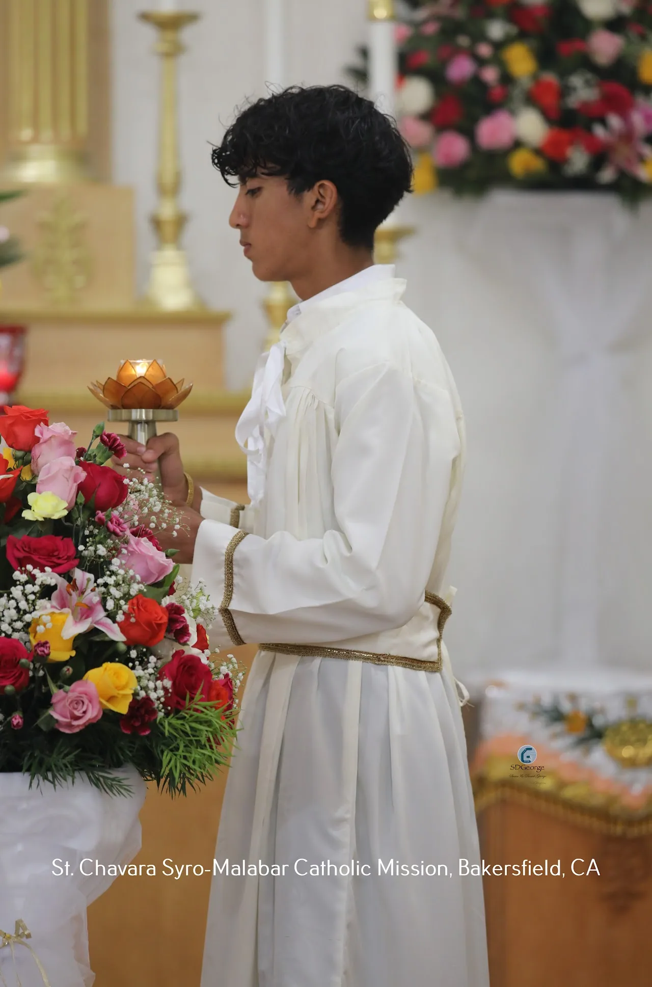 Altar server in white alb holding a lotus-shaped brass lamp before the gilded altar