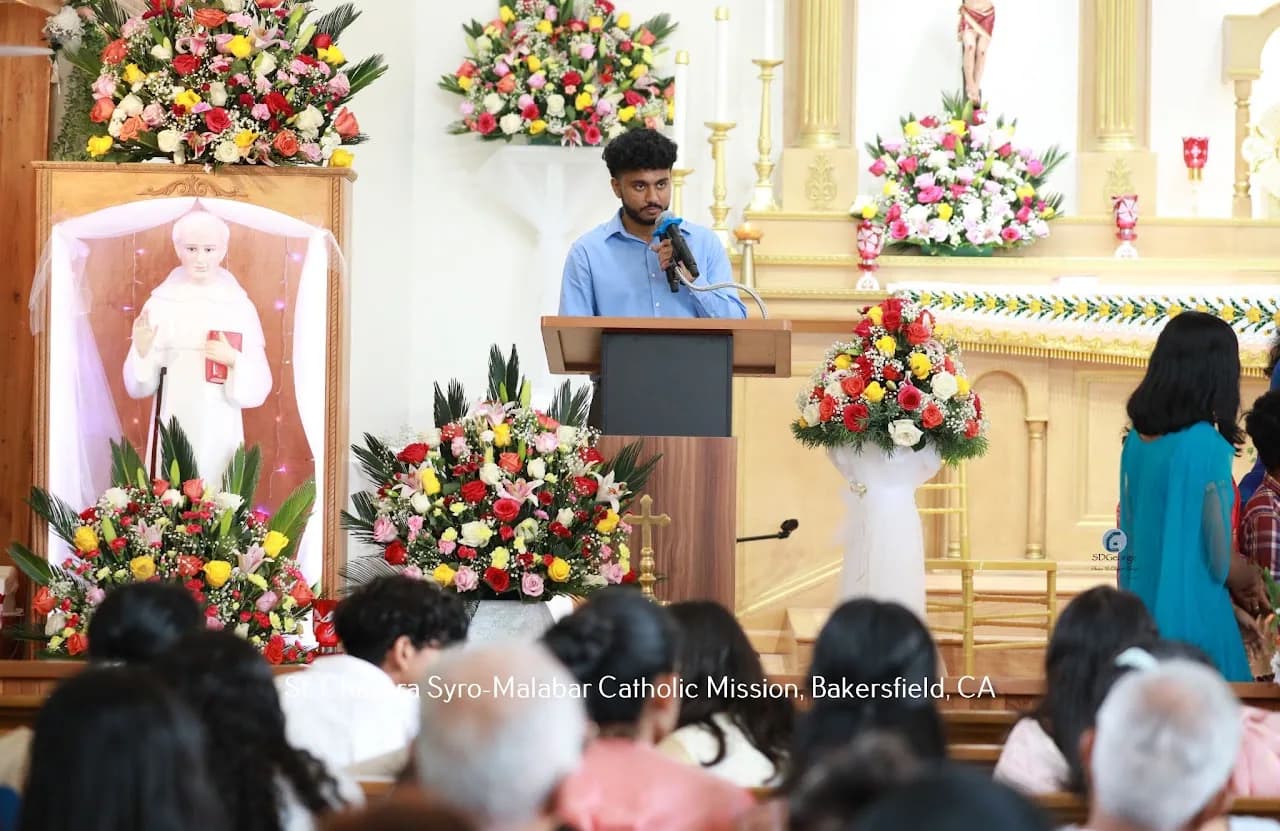 Young man speaking at the lectern beside the flower-decked St. Chavara shrine during a parish celebration