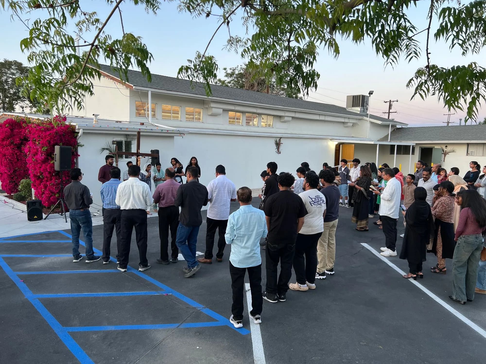 Parishioners gathered in a semicircle at dusk before an outdoor crucifix on the parish hall wall