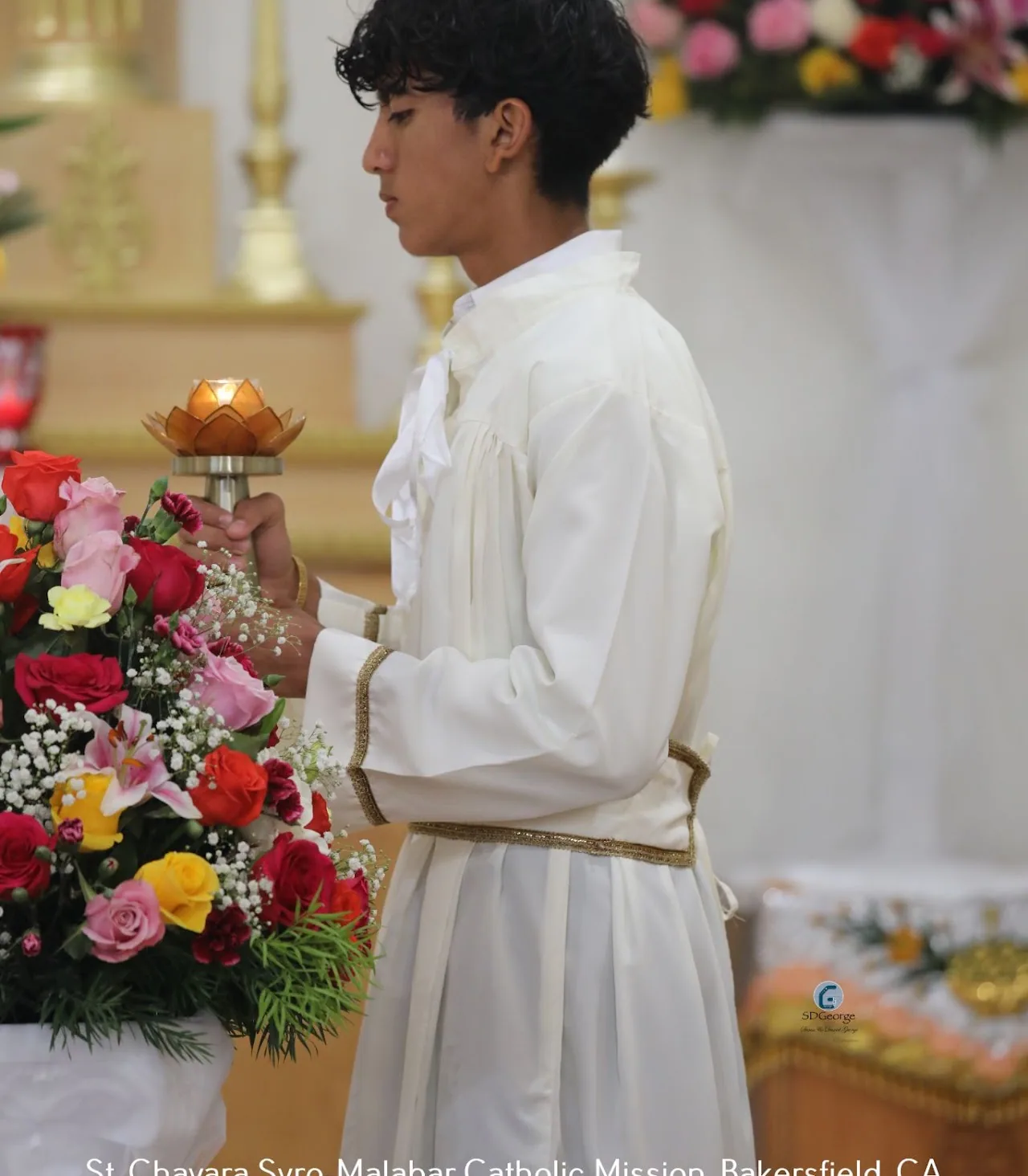 Altar server in a gold-trimmed white alb lifting a lotus-shaped brass lamp by the altar flowers