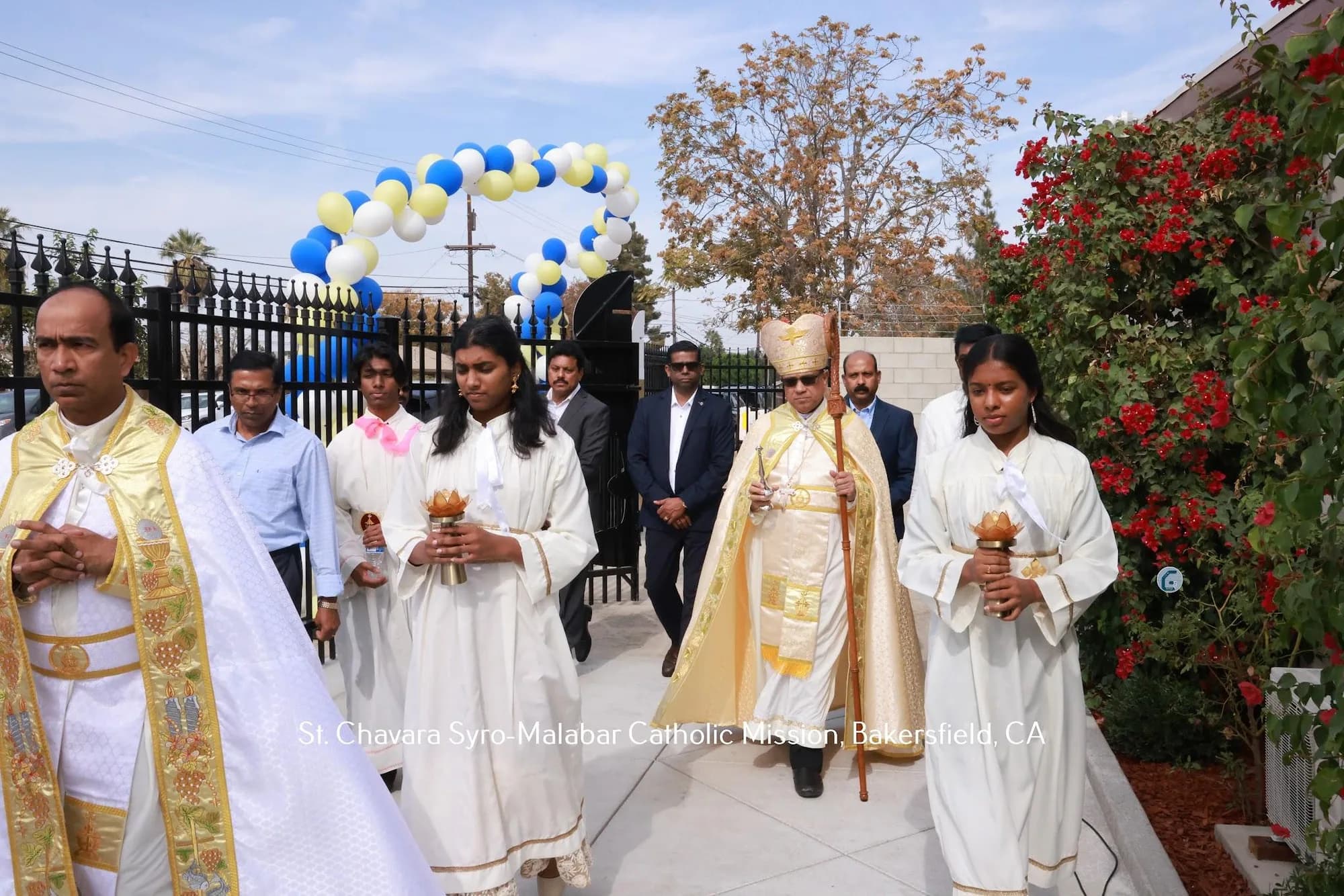 Bishop with crozier processing behind two altar servers carrying brass lamps, under a balloon arch