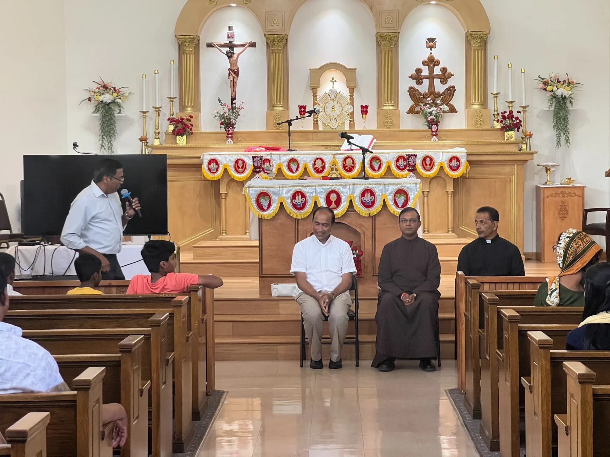 Man speaking into a microphone at a parish meeting while three seated leaders and families listen from the pews