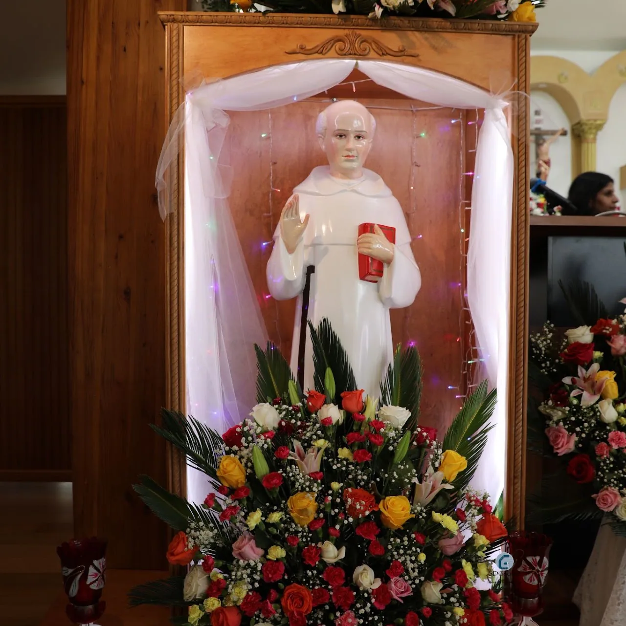 White statue of St. Chavara in a wooden shrine niche framed by tulle, fairy lights and a bank of flowers