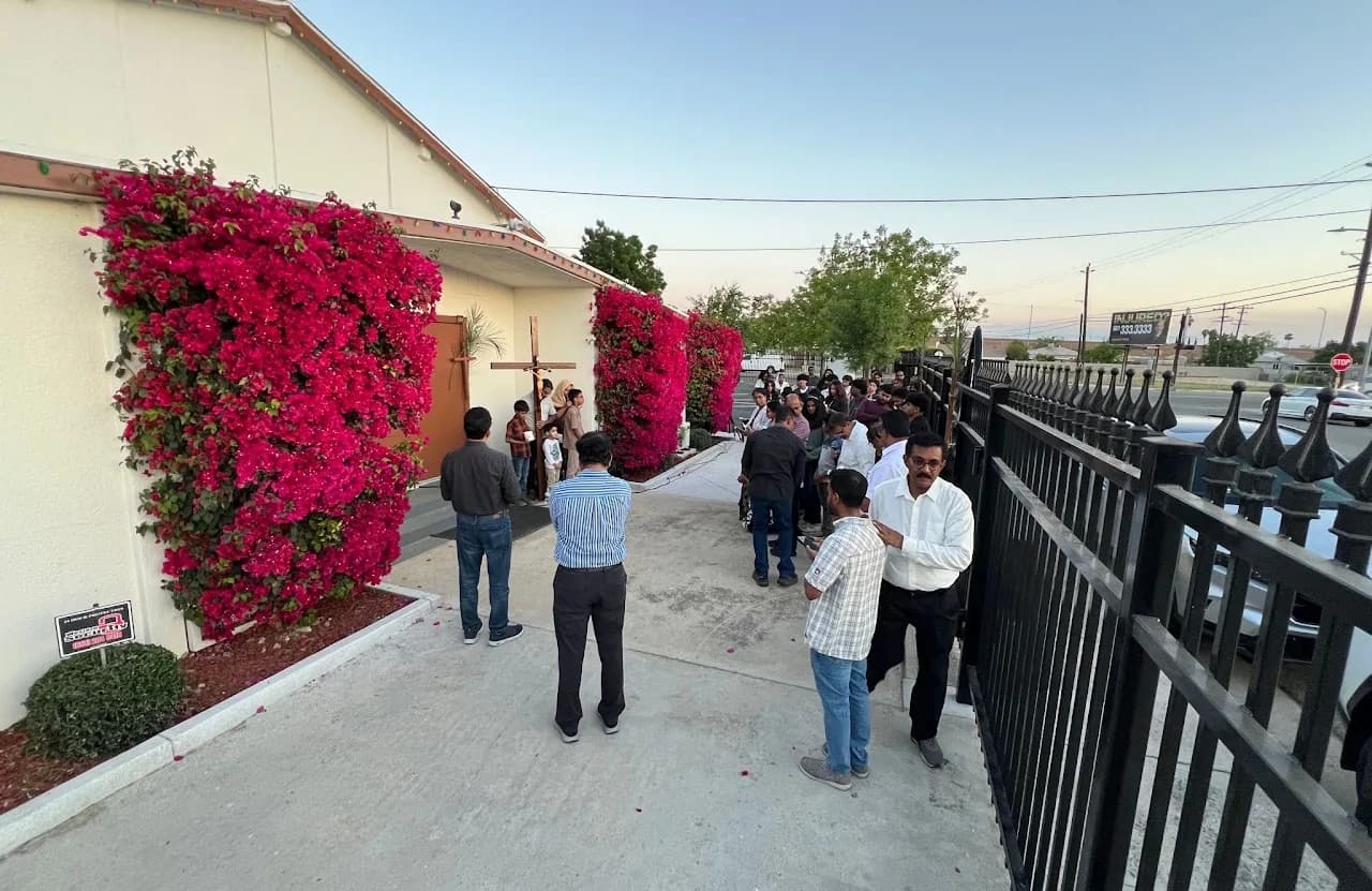 Evening Holy Week gathering outside the church, crowd beside bougainvillea walls as a wooden cross is carried in