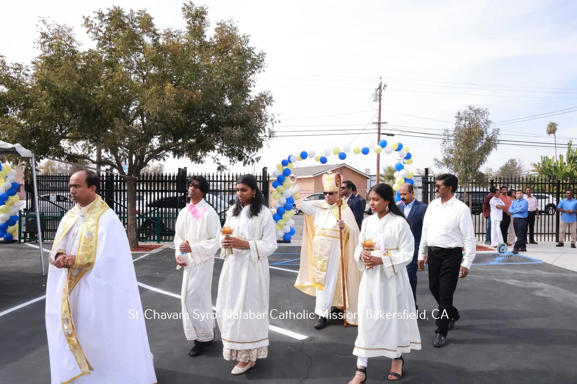 Bishop blessing during an outdoor procession behind altar servers with brass lamps, under a balloon arch