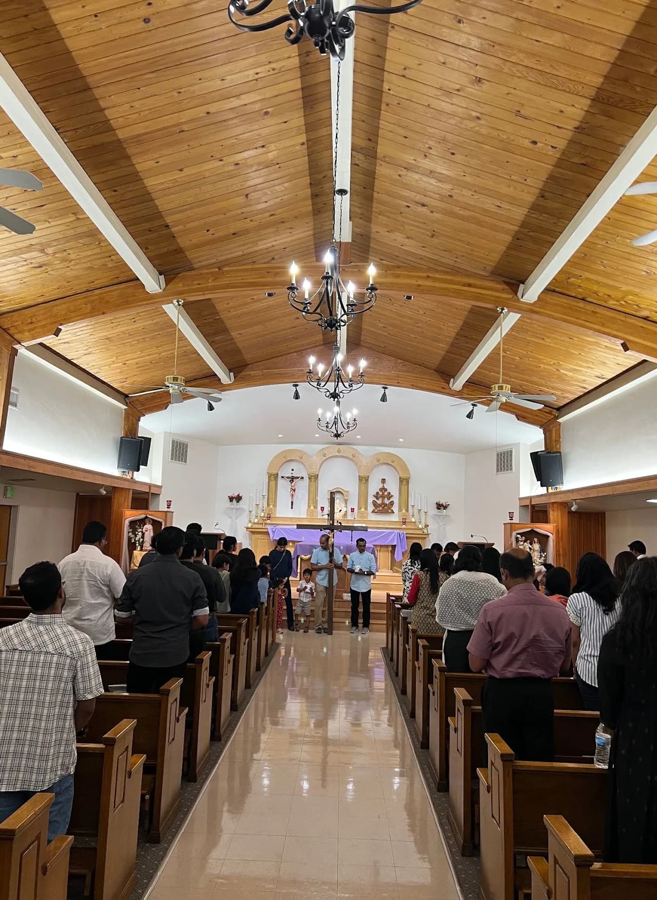 Congregation standing in wooden-ceilinged nave as men carry a large wooden cross up the center aisle