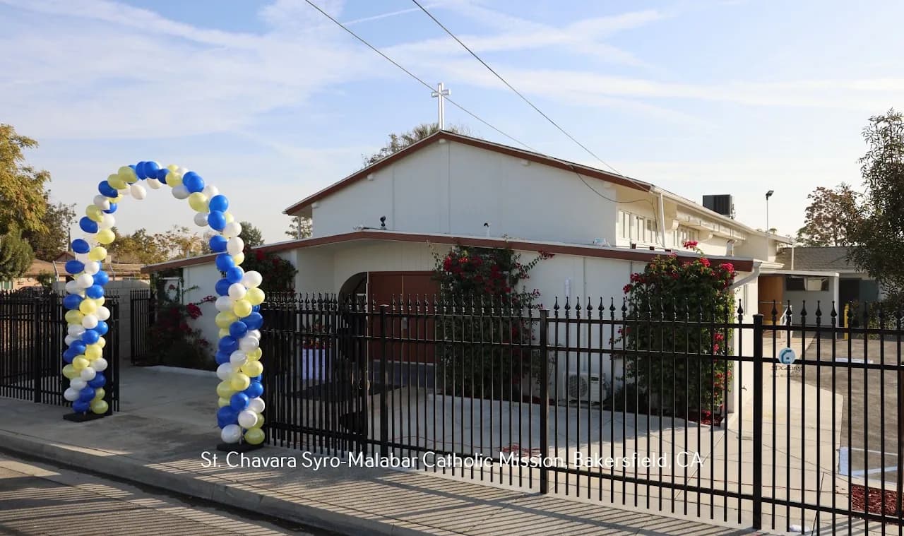 White church with rooftop cross behind an iron fence, blue and yellow balloon arch at the entrance