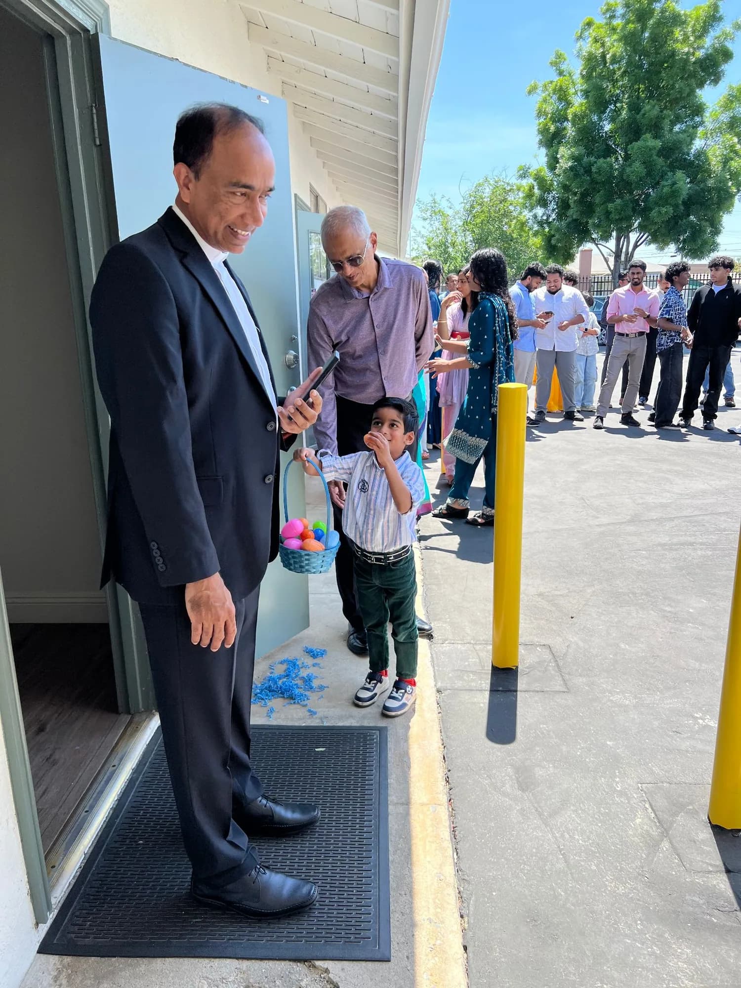 Priest in doorway smiling at a boy holding a basket of colored Easter eggs outside the church