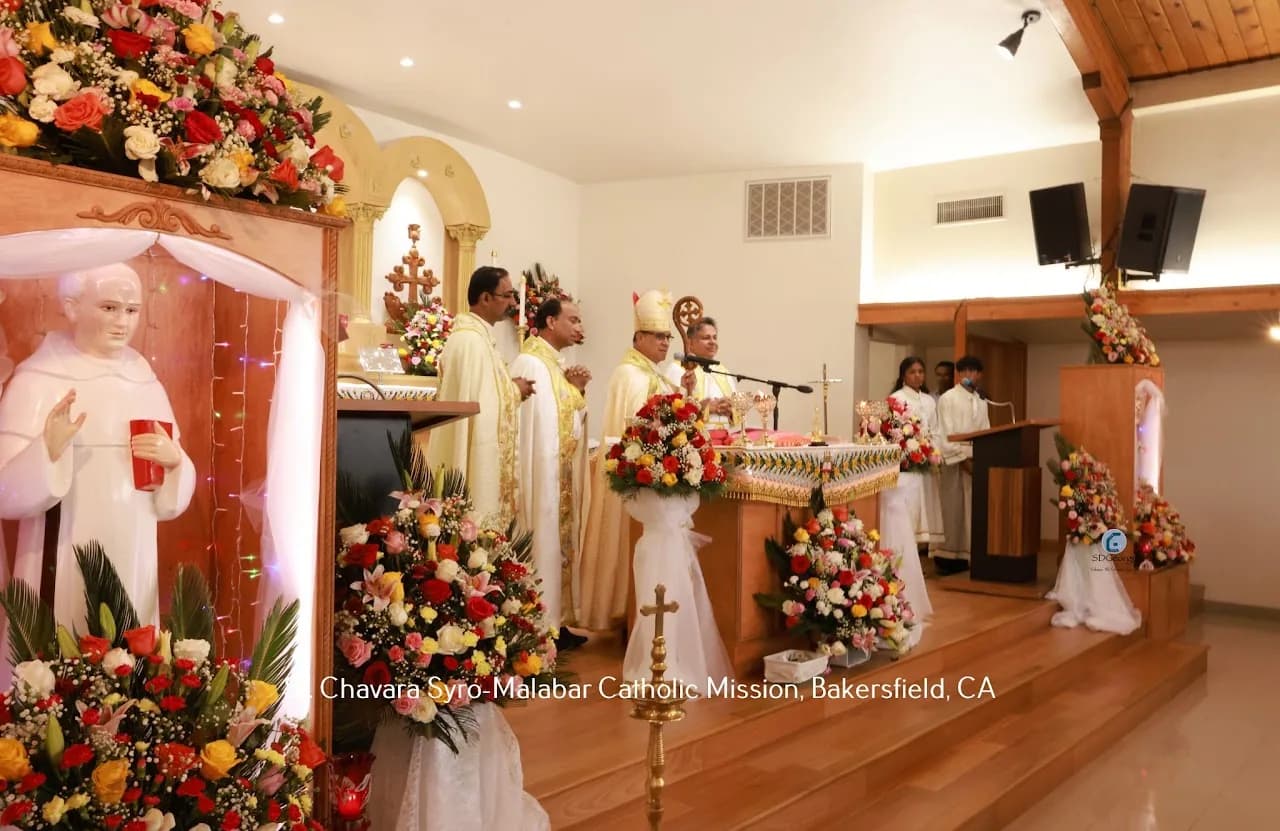 Bishop and concelebrating priests at a garlanded altar beside an illuminated St. Chavara shrine