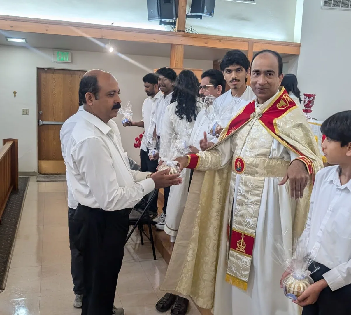 Priest in vestments handing a cellophane-wrapped gift to a man as others wait in line
