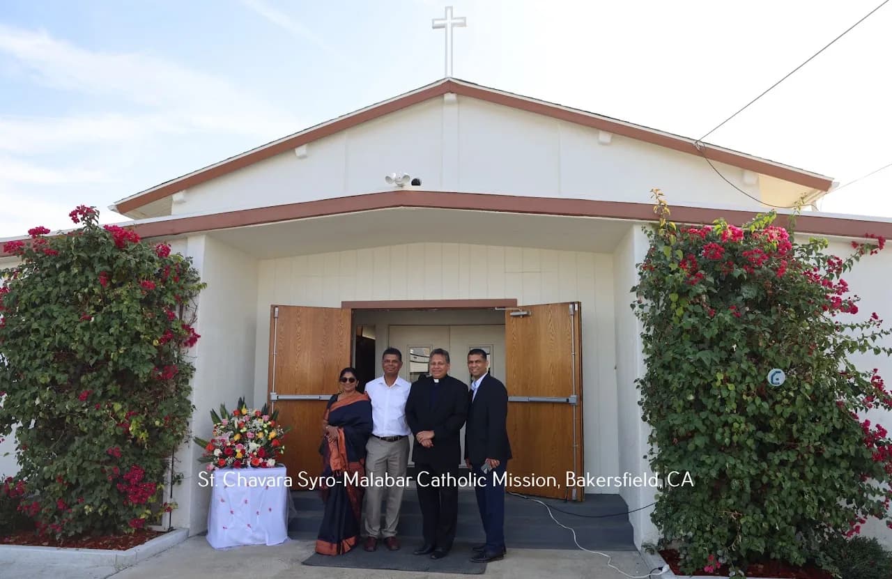 Four people including a priest standing at the church entrance framed by flowering bougainvillea