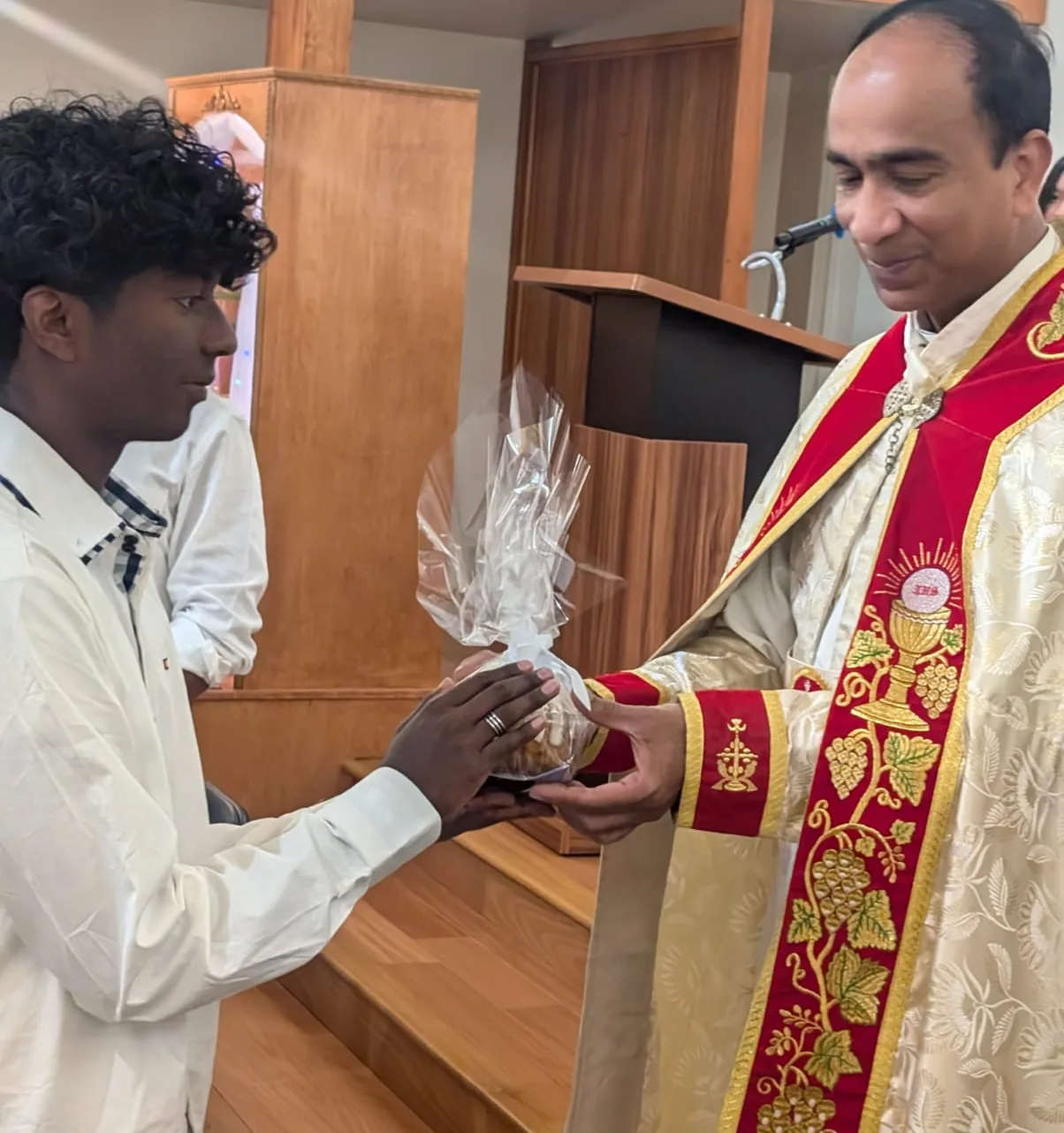 Priest in red and cream vestments handing a wrapped gift basket to a young man