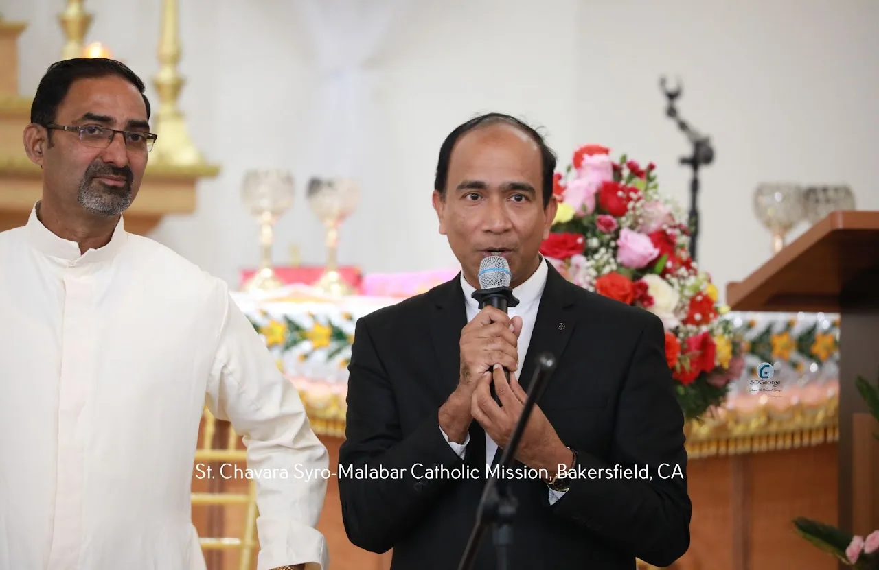 Man in a suit addressing the congregation on a microphone as a priest stands beside him