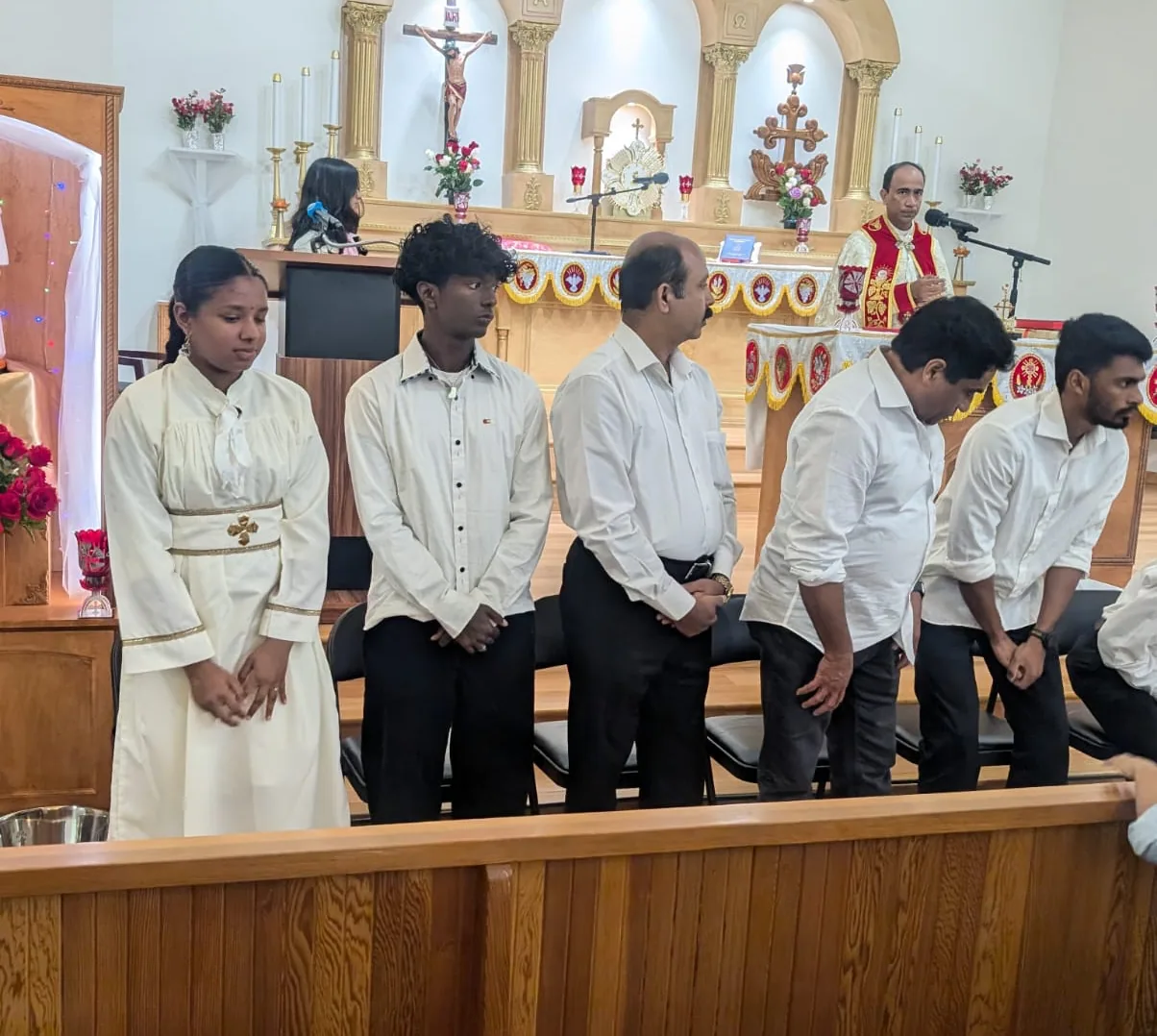 Six parishioners standing with bowed heads in the sanctuary as a priest in red speaks
