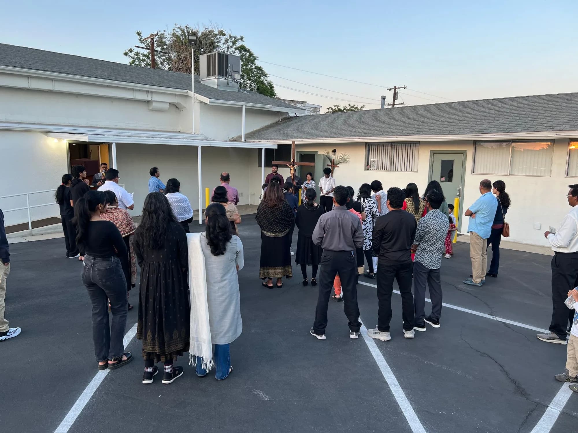 Parishioners gathered in the parking lot at dusk around a large wooden crucifix during an outdoor prayer service