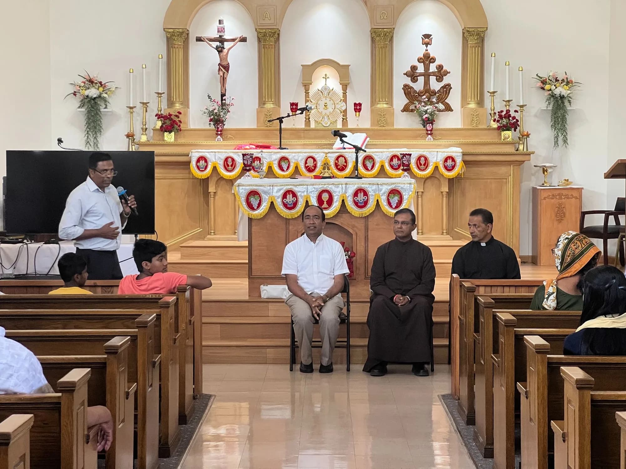 Man speaking into a microphone in the sanctuary while three seated men, two in clerical dress, listen