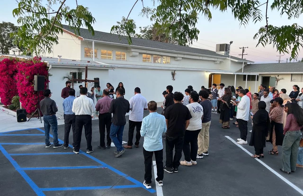About forty parishioners standing in a parking lot at dusk facing a wooden cross beside blooming bougainvillea