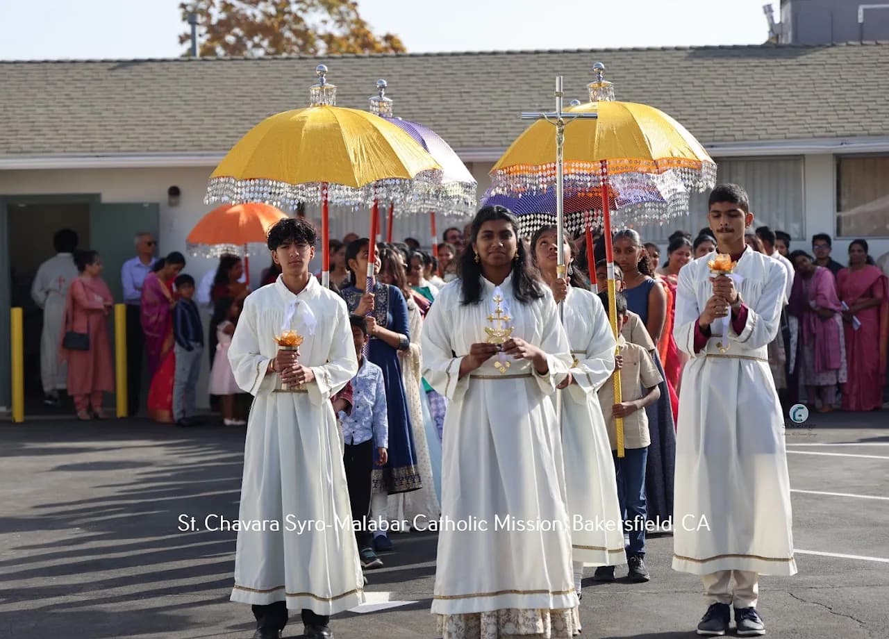 Altar servers in white robes lead an outdoor procession under yellow and orange Kerala ceremonial umbrellas