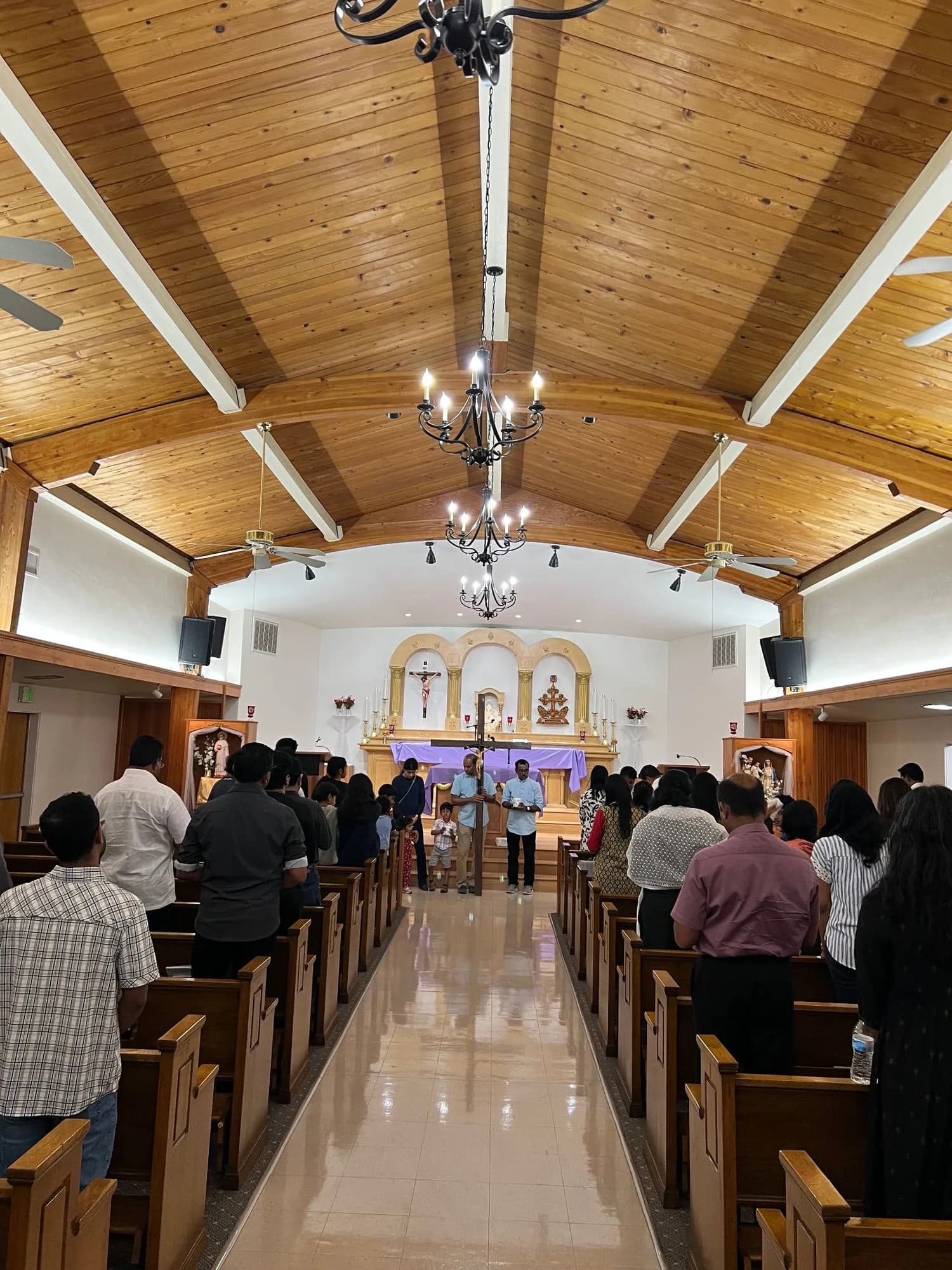 Congregation standing in pews under a wooden vaulted ceiling as two men hold a wooden cross by the purple-draped altar
