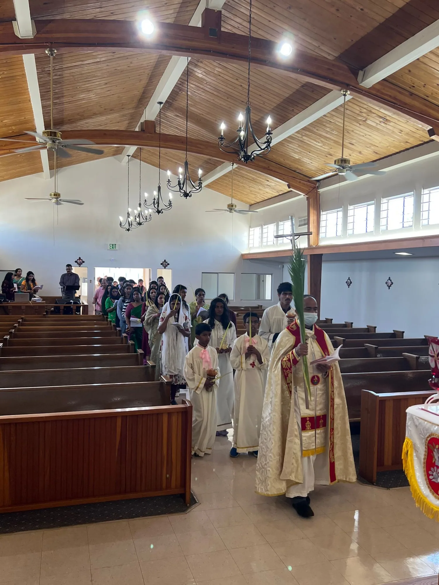 Priest in cream and red vestments carries a palm up the aisle behind a processional cross and two altar servers