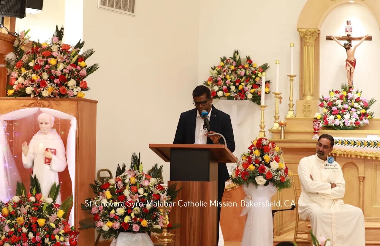 Man in a suit speaking at a lectern in the flower-decorated sanctuary while a vested priest sits nearby