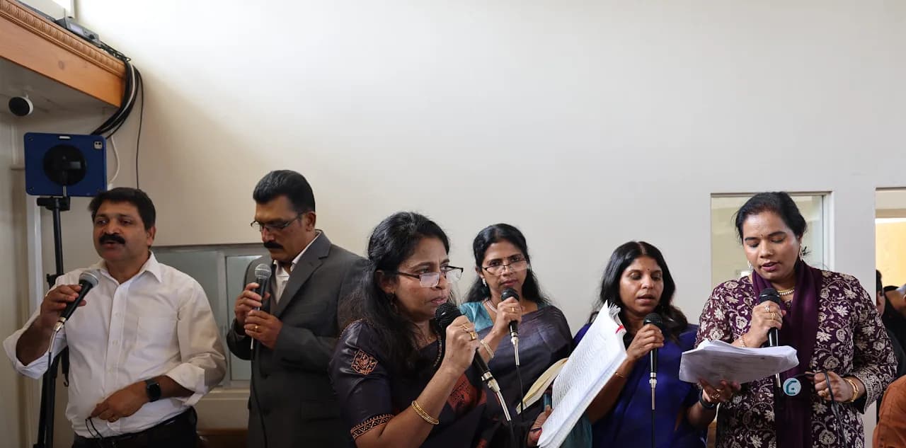 Six adult choir members singing into microphones while holding sheet music in a parish hall