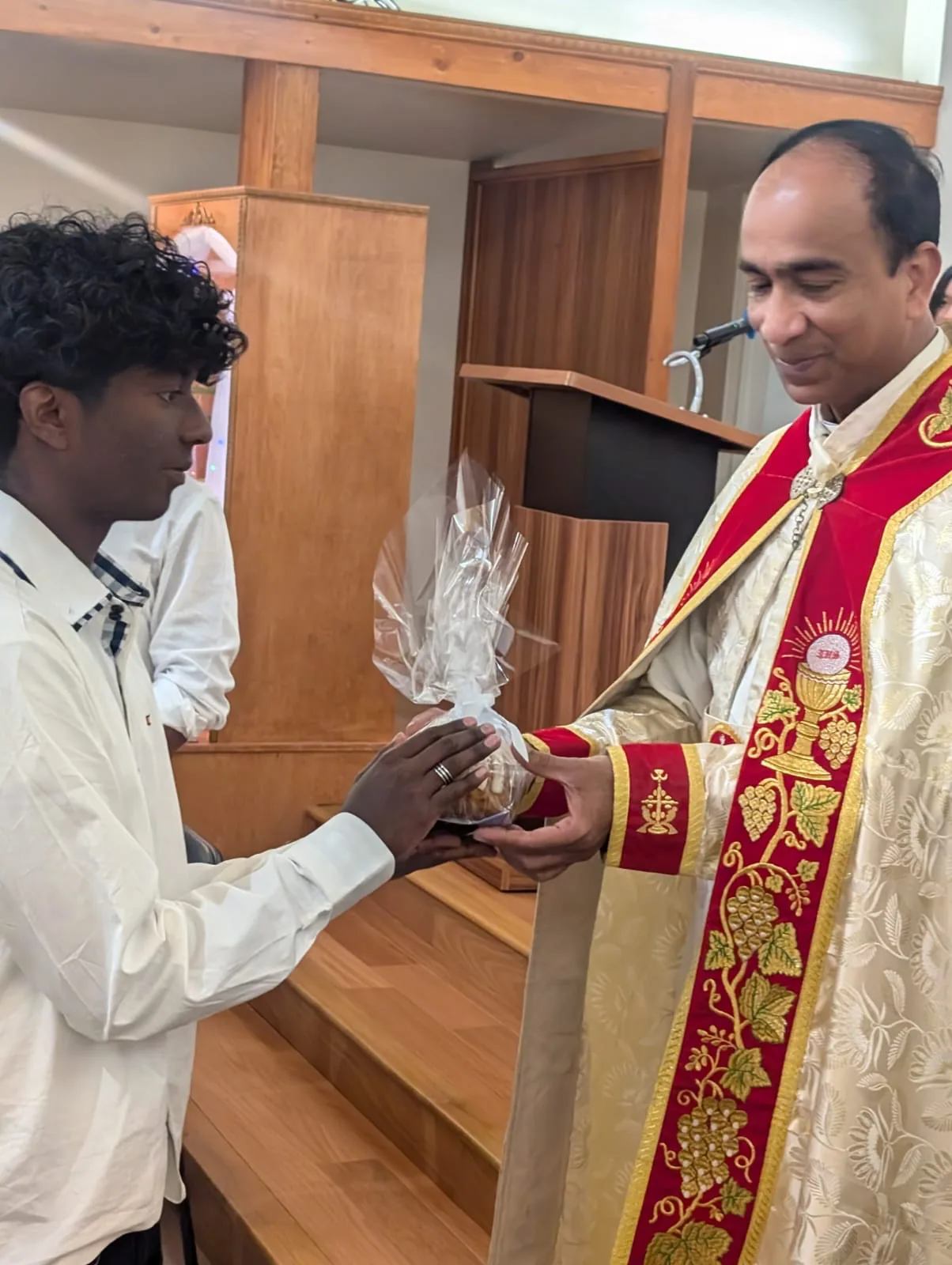 Priest in red and gold vestments handing a wrapped gift basket to a young man on the sanctuary steps