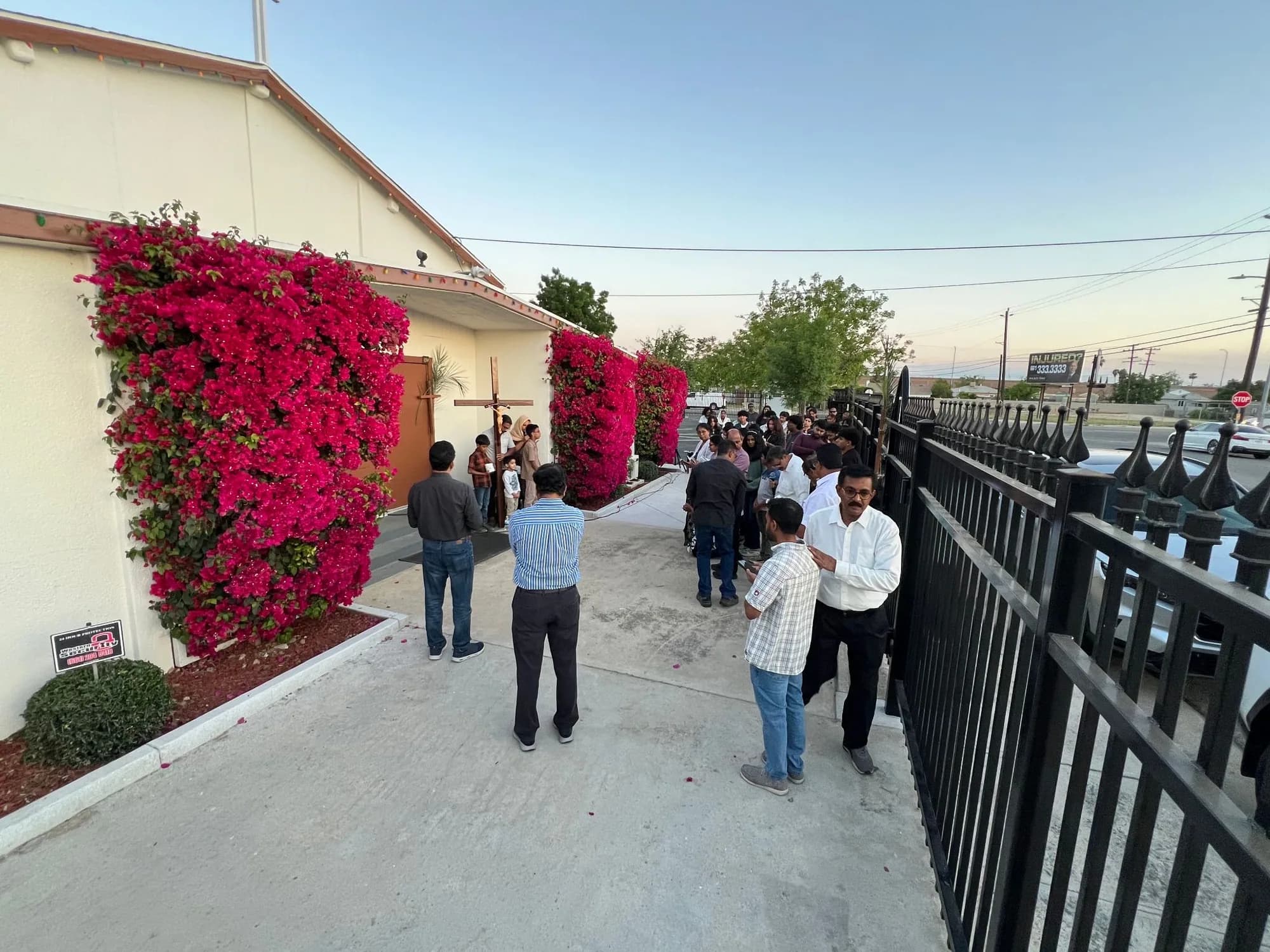 Parishioners gathered at dusk beside bougainvillea-covered church walls as a wooden crucifix is carried